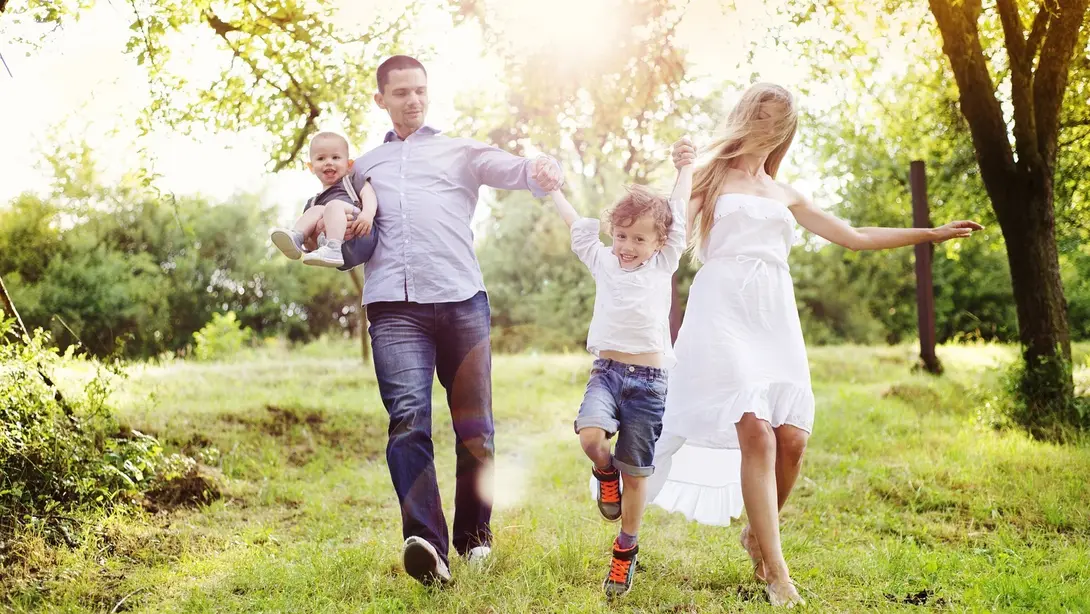 Familienfreundlich wohnen und Haus bauen im Grünen Familie mit zwei Kindern läuft bei Sonnenschein durch eine grüne Wiese in naturnaher UmgebungFamilienfreundlich wohnen und Haus bauen im Grünen