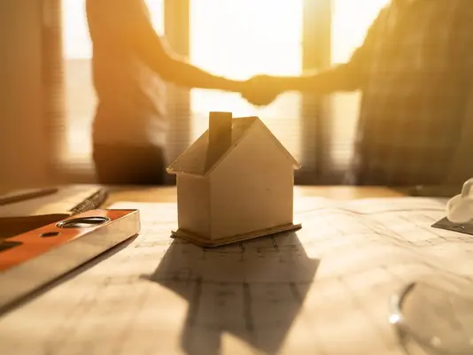 Modellhaus auf Bauunterlagen vor zwei Personen, die sich die Hand geben