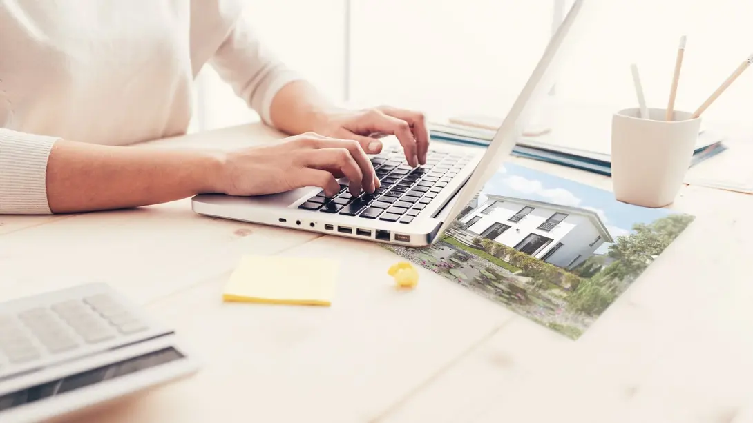 Hausbau online planen und Beratung anfragen Person plant am Laptop den Hausbau mit Hausbild und Unterlagen auf dem Schreibtisch