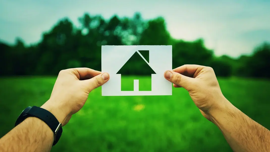 Hände halten ein Haus-Symbol vor einer grünen Wiese als Symbol für Grundstückssuche und Eigenheimplanung