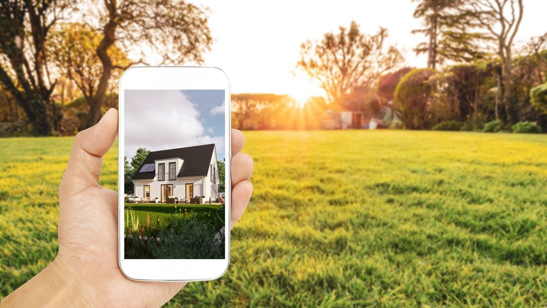 Grundstück finden und Hausbau in Leese planen Hand hält Smartphone mit Hausmotiv vor einer grünen Grundstücksfläche als Symbol für Grundstückssuche und Hausbau in Leese mit Contura Bau GmbH