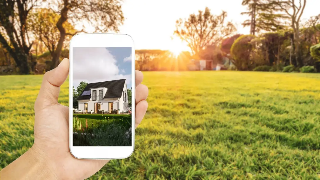 Digitale Hausplanung auf dem Grundstück Smartphone mit Einfamilienhaus-Visualisierung vor einer freien Grünfläche als Symbol für digitale Grundstücks- und Hausplanung