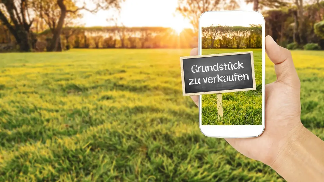 Baugrundstück zum Verkauf finden Hand hält ein Smartphone mit einem Verkaufsschild vor einem freien Grundstück