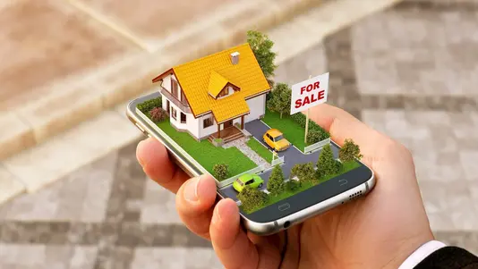 Grundstücks- und Immobiliensuche in Bassum-Neubruchhausen Hand hält ein Smartphone mit Hausmodell und Verkaufsschild als Symbol für Grundstückssuche und Immobilienkauf in Bassum-Neubruchhausen.