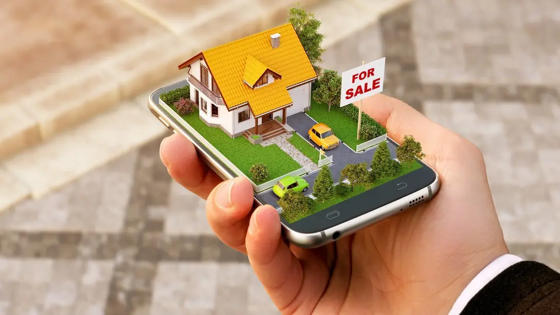 Hand hält ein Smartphone mit Hausmodell und Verkaufsschild als Symbol für Grundstückssuche und Immobilienkauf in Diepenau-Essern.