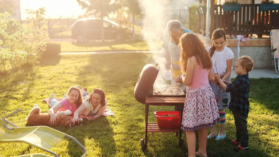 Mehrgenerationen-Familie beim Grillen im Garten mit Kindern, Großvater und Haustieren bei Abendsonne