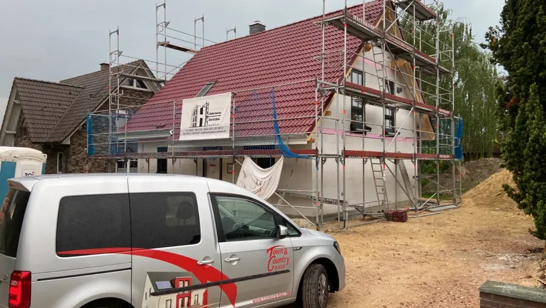 Einfamilienhaus im Bau mit Gerüst, rotem Dach und Town & Country Haus Fahrzeug auf der Baustelle