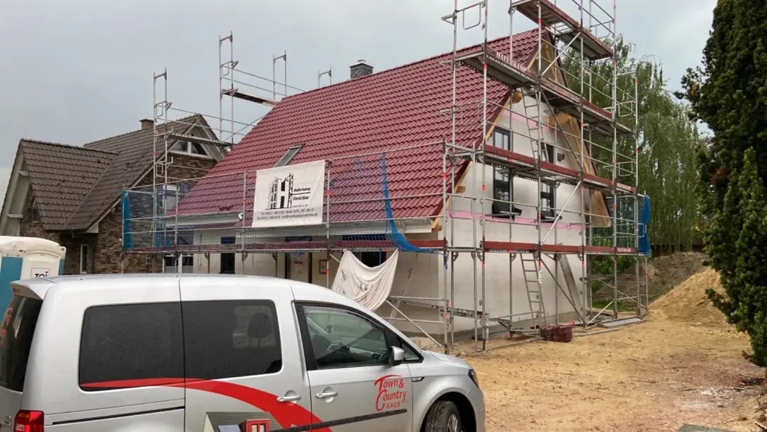 Einfamilienhaus im Bau mit Gerüst, rotem Dach und Town & Country Haus Fahrzeug auf der Baustelle